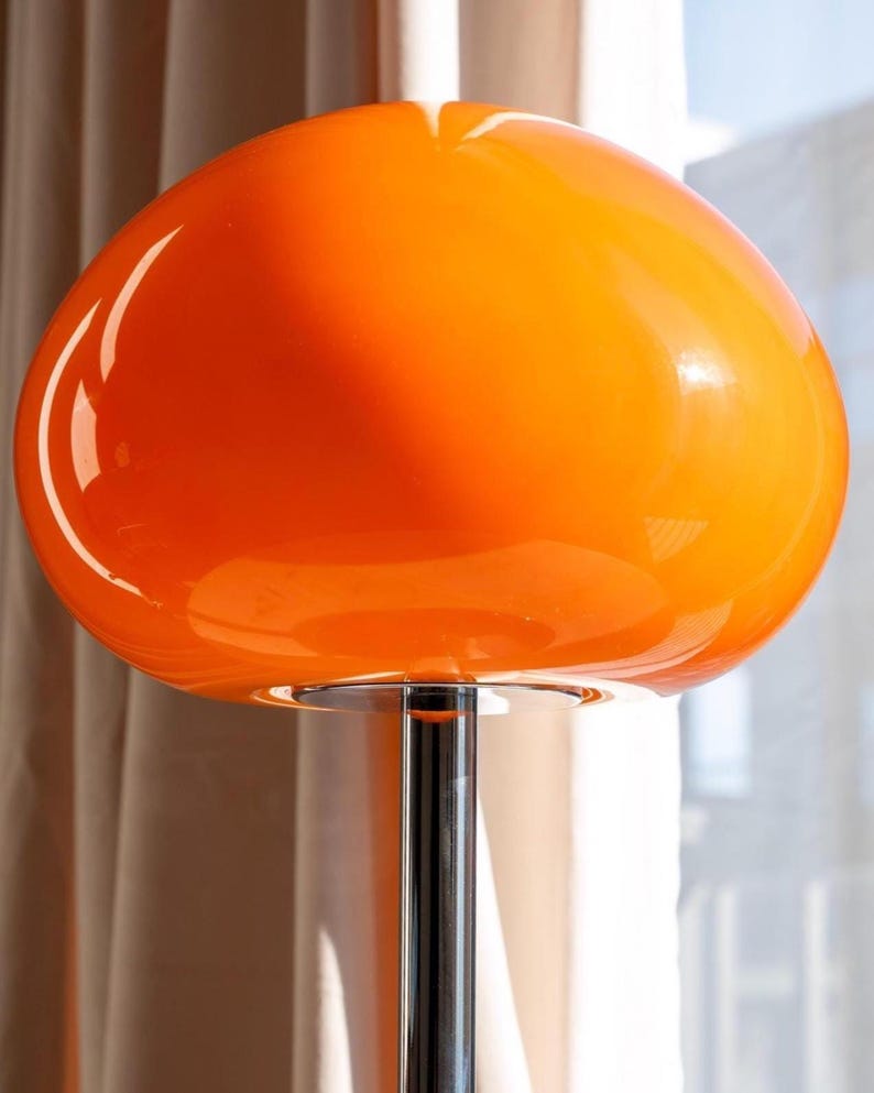 May include: A vibrant orange, mid-century modern style lamp with a large, rounded shade. The shade sits atop a chrome-colored, slender pole. The lamp's design is reminiscent of a mushroom shape, casting a warm glow.