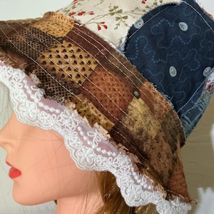 May include: A patchwork bucket hat with a white lace brim. The hat features various fabric patterns in shades of brown, beige, and navy blue, with floral and dotted designs. The brim is trimmed with delicate white lace.