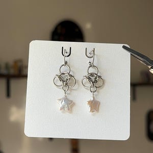 Könnte beinhalten: Ein Paar silberfarbene Ohrringe mit einem kreisförmigen Metallglied-Design im Kettengeflecht-Stil, an dem ein kleiner, schimmernder, sternförmiger Perlenanhänger befestigt ist. Die Ohrringe sind auf einer weißen Karte befestigt.