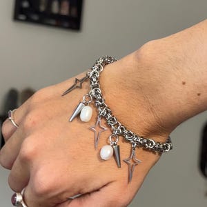Könnte beinhalten: Ein silberfarbenes Kettenarmband mit hängenden Anhängern, darunter mehrere vierzackige Sterne, kleine Metallspitzen und glatte weiße ovale Perlen. Das Armband besteht aus ineinandergreifenden Metallringen in einem dichten Muster.