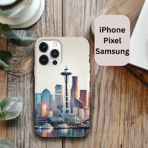 May include: A white phone case with a colorful image of the Seattle skyline, including the Space Needle. The case has a camera lens cutout and the words "iPhone Pixel Samsung" are printed on a separate pink rectangle.