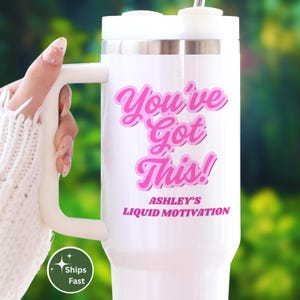 May include: White travel mug with a handle, featuring the text "You've Got This!" in pink script. The mug also includes the text "Ashley's Liquid Motivation" in pink. The mug is held by a person wearing a white sweater.
