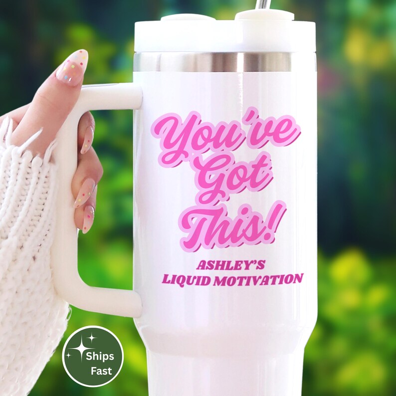 May include: White travel mug with a handle, featuring the text "You've Got This!" in pink script. The mug also includes the text "Ashley's Liquid Motivation" in pink. The mug is held by a person wearing a white sweater.