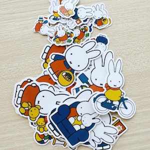 Op de afbeelding: Een verzameling stickers met een wit konijn met zwarte contouren. De stickers tonen het konijn in verschillende outfits en activiteiten, waaronder fietsen. De stickers zijn in oranje, geel en blauw.