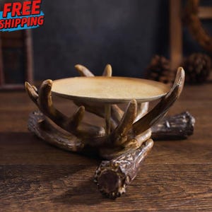 May include: A decorative candle holder with a rustic antler design. The holder features a round, light-colored tray supported by a detailed antler base. The antlers are brown with textured details, creating a natural aesthetic.
