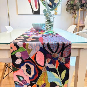 以下が含まれることがあります： ピンク、ブルー、グリーン、イエロー、ブラックの色の花柄が施されたカラフルなテーブルランナー。ランナーは布製で、モダンで抽象的なデザインです。