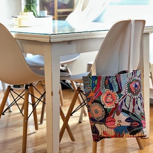 Puede incluir: Una bolsa de tela colorida con un patr&oacute;n floral y geom&eacute;trico cuelga de una silla blanca. La bolsa tiene una correa de lona blanca.