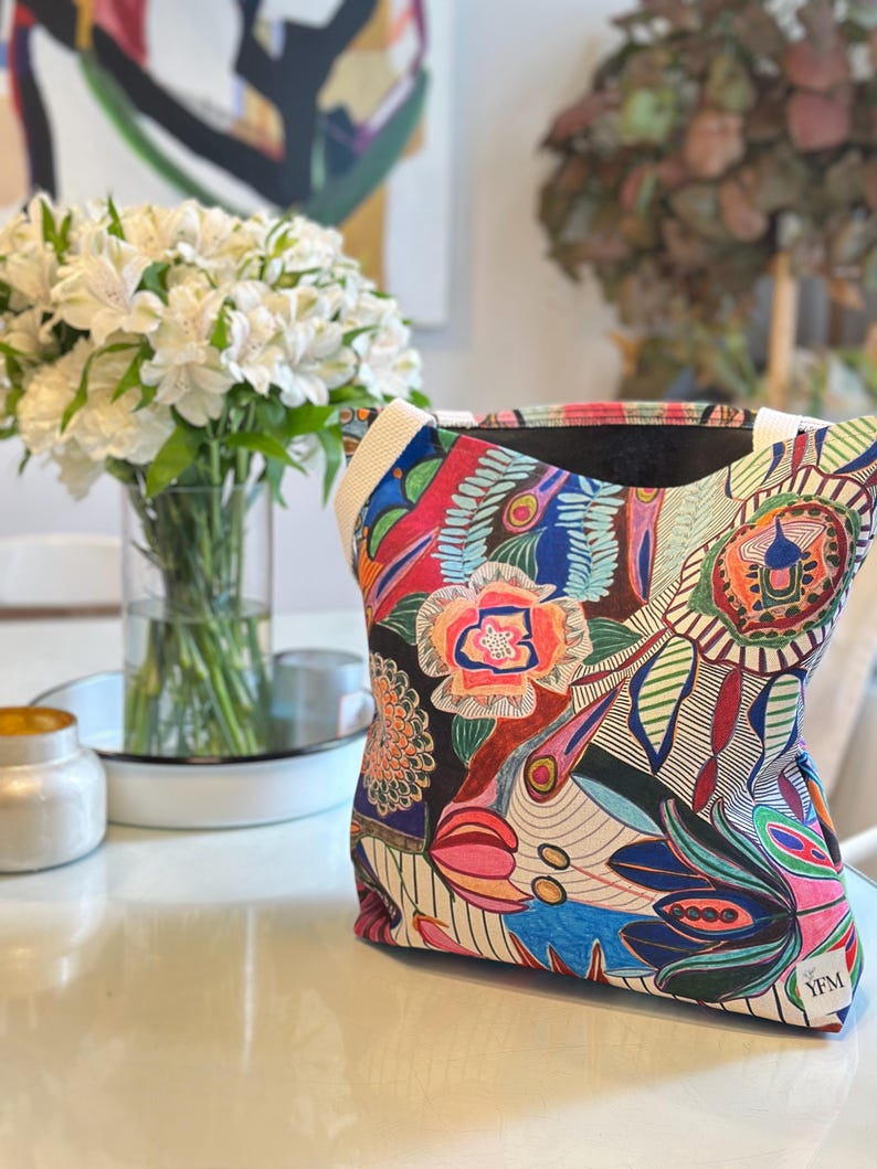 Puede incluir: Una bolsa de tela colorida con un dise&ntilde;o floral y abstracto. La bolsa tiene un forro negro y una correa blanca. La bolsa tiene el texto "YFM" en la esquina inferior derecha.