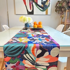 May include: A colourful table runner with a modern abstract design featuring a mix of bright colours, including red, orange, yellow, green, blue, and purple. The runner is made of a soft, silky fabric and has a geometric pattern.