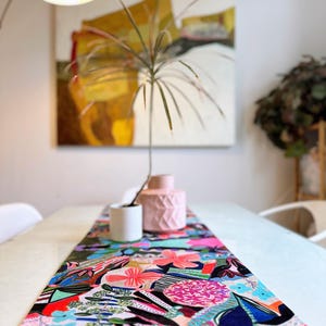 May include: A colorful floral table runner with a vibrant design of pink, blue, and purple flowers. The runner is placed on a white table, with a pink vase and a white planter. A painting hangs in the background.