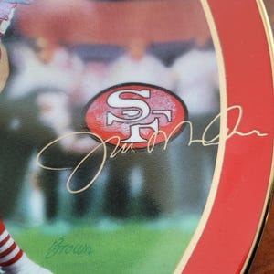 May include: A red and white ceramic plate with a gold rim features a photo of a football player wearing a red and white uniform. The player's signature is written in gold script, along with the San Francisco 49ers logo.