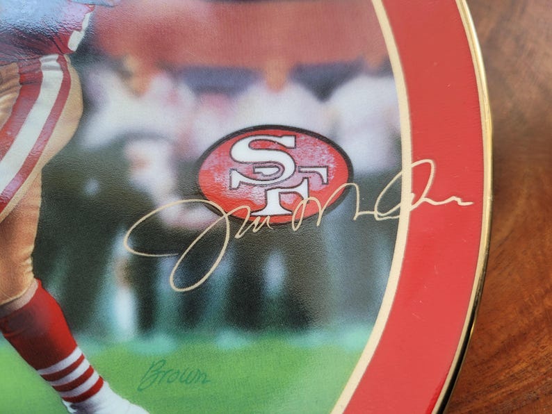 May include: A red and white ceramic plate with a gold rim features a photo of a football player wearing a red and white uniform. The player's signature is written in gold script, along with the San Francisco 49ers logo.