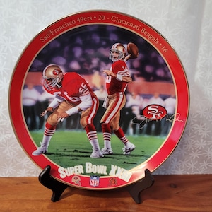 May include: A red and gold ceramic plate with a picture of two football players in red and gold uniforms. The plate commemorates Super Bowl XXIII, with the text "San Francisco 49ers - 20 - Cincinnati Bengals - 16" and "Super Bowl XXIII" printed on the plate.