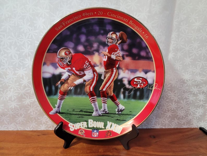May include: A red and gold ceramic plate with a picture of two football players in red and gold uniforms. The plate commemorates Super Bowl XXIII, with the text "San Francisco 49ers - 20 - Cincinnati Bengals - 16" and "Super Bowl XXIII" printed on the plate.