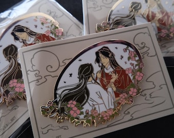 Op de afbeelding: Gouden emaille pin met twee figuren met lang zwart haar, een in een witte jurk en de ander in een rode jurk, omringd door roze bloemen op een witte achtergrond.