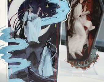Op de afbeelding: Acryl standees met anime-stijl illustraties. Eén toont een figuur in witte gewaden op een halve maan met blauwe accenten, de andere toont twee figuren die elkaar omarmen in een doodskistvormig frame.