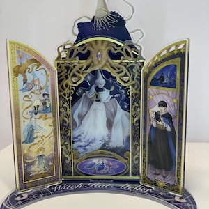 May include: A multi-panel acrylic display stand featuring Witch Hat Atelier artwork. The central panel shows a character in a white robe under a golden tree structure, flanked by two side panels with various character illustrations and gold accents.