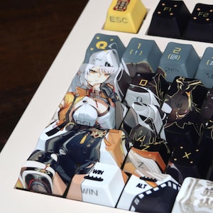 May include: A close-up of a white mechanical keyboard with custom keycaps. The keycaps feature anime-style illustrations and various colors, including yellow, black, and blue. The keyboard has a white frame and a wooden surface.