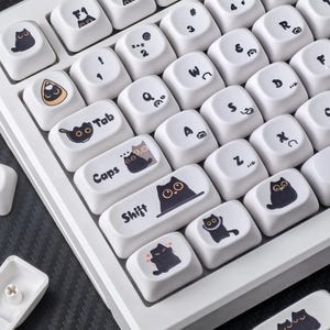 Op de afbeelding: Wit mechanisch toetsenbord met aangepaste keycaps met zwarte kattenillustraties. De keycaps hebben verschillende symbolen en tekst, waaronder "Tab", "Caps" en "Shift". De spatiebalk leest "Having a Good Time".