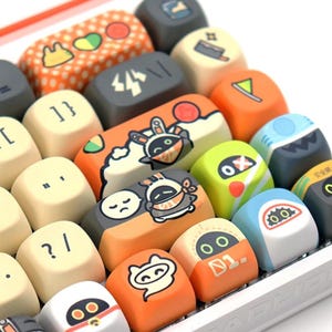 May include: A close-up of a mechanical keyboard with custom keycaps. The keycaps feature various cartoon designs, including a rabbit, a shark, and other colorful graphics. The keycaps are in shades of orange, white, gray, and green.