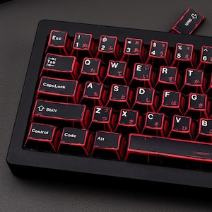 May include: Black mechanical keyboard with red backlit keys. The keys have white lettering and Japanese characters. The keyboard includes keys for Shift, Control, Alt, and Caps Lock. The keyboard is on a dark surface.