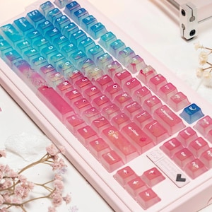 May include: A pink mechanical keyboard with gradient keycaps transitioning from blue to pink. The keyboard has a light pink frame and a standard QWERTY layout. The keycaps are translucent, and the keyboard is surrounded by decorative flowers.