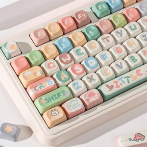 May include: A pastel-coloured keyboard with custom keycaps. The keys feature letters, numbers, and symbols in various colours, along with decorative icons. The keyboard includes the text "LITTLE FORES".