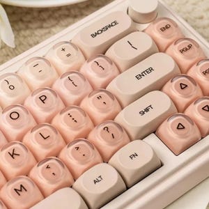 Puede incluir: Un teclado de estilo retro con teclas redondas de color melocotón y un marco beige claro. Las teclas presentan letras y símbolos blancos, incluyendo "BACKSPACE", "ENTER" y "SHIFT". El teclado tiene un diseño vintage.
