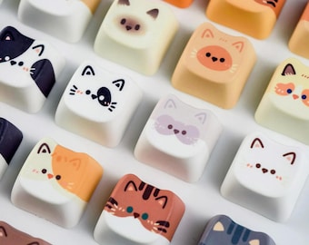 May include: A collection of colorful keyboard keycaps featuring various cat face designs. The keycaps have a white base with orange, brown, gray, and black cat faces. Each keycap has a unique cat expression.