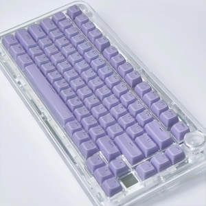 May include: A clear, rectangular keyboard with lavender keycaps. The keyboard has a standard layout with function keys and a number pad. The keycaps are a solid lavender color, and the keyboard has a clear, transparent frame.