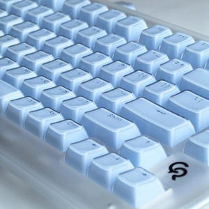Puede incluir: Primer plano de un teclado mec&aacute;nico azul claro. El teclado tiene una distribuci&oacute;n est&aacute;ndar con teclas azul claro. Las teclas est&aacute;n dispuestas en filas y columnas, con la tecla Shift en un lugar destacado. El teclado tiene un marco de pl&aacute;stico transparente.