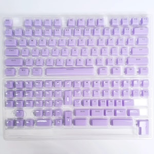 May include: Two keyboard covers with a light purple keycap design. The keycaps are arranged in a standard QWERTY layout, with alphanumeric keys, function keys, and a number pad. The covers are made of a clear material.