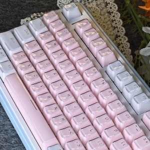 May include: A close-up of a mechanical keyboard with a light blue frame and white and pink keycaps. The keycaps are arranged in a standard QWERTY layout. The keyboard is on a gray surface.