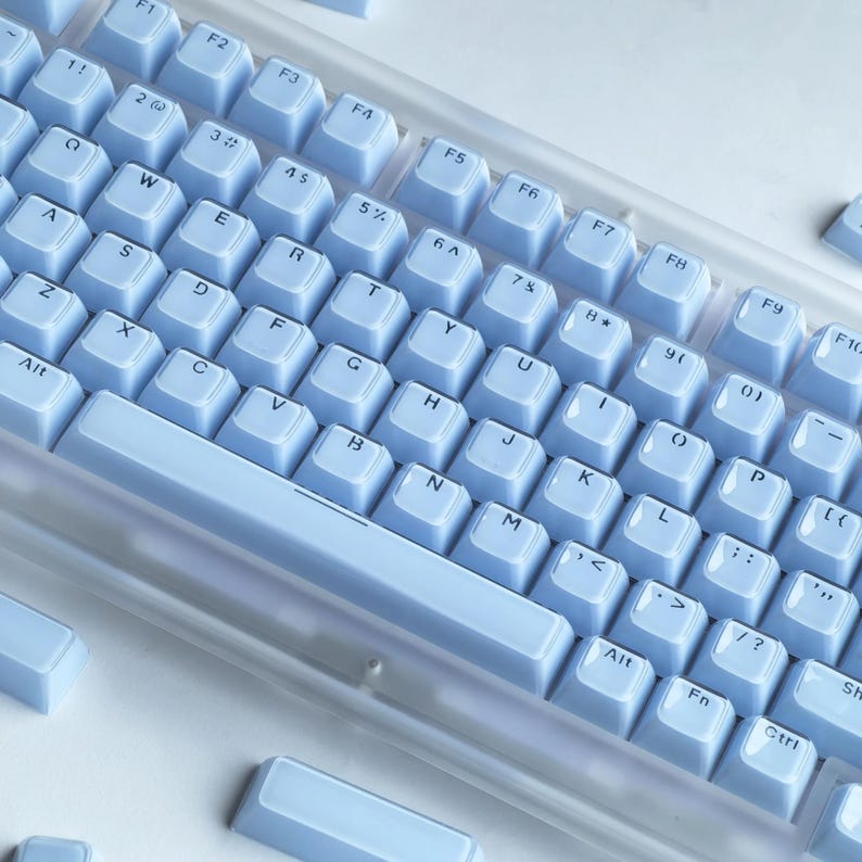 Puede incluir: Un teclado mec&aacute;nico azul claro con letras blancas. El teclado tiene un marco de pl&aacute;stico transparente y est&aacute; sobre un fondo blanco. Las teclas est&aacute;n dispuestas en una distribuci&oacute;n QWERTY est&aacute;ndar.