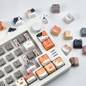 K&ouml;nnte beinhalten: Eine Sammlung von Tastenkappen im Katzen-Design in verschiedenen Farben, darunter Orange, Wei&szlig;, Grau und Schwarz. Die Tastenkappen sind auf einer wei&szlig;en Tastaturbasis angeordnet, einige liegen lose herum. Die Katzen haben unterschiedliche Gesichter.