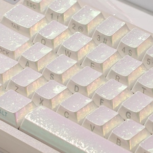 May include: A computer keyboard featuring white keycaps with a shimmering, iridescent finish that reflects rainbow colours. The keys display standard alphanumeric characters and symbols, including ESC, TAB, and ALT, in a clear, dark grey font.
