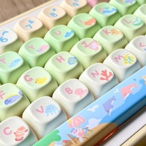 K&ouml;nnte beinhalten: Nahaufnahme einer Tastatur mit individuellen Tastenkappen. Die Tastenkappen sind in Pastellfarben gehalten, darunter Gr&uuml;n, Creme und Gelb. Jede Tastenkappe zeigt einen Buchstaben oder eine Zahl und eine kleine, farbenfrohe Illustration von Meeresbewohnern. Die Leertaste hat eine Strandszene.