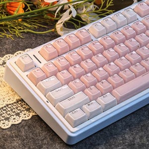 May include: A white and light blue mechanical keyboard with pink keycaps. The keyboard has a standard layout with function keys and a number pad. The keycaps are a soft pink color, and the keyboard has a clean, modern design.