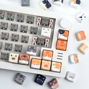K&ouml;nnte beinhalten: Eine wei&szlig;e Tastatur mit Keycaps im Katzen-Design. Die Keycaps zeigen verschiedene Katzen-Gesichter in Farben wie Orange, Grau und Braun. Die Tastatur hat auch transparente mechanische Schalter und eine wei&szlig;e runde Taste.