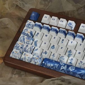 May include: A mechanical keyboard with a dark wood frame and white keycaps featuring blue floral and bird patterns. The keys include standard QWERTY lettering, function keys labeled F1 through F5, and a long spacebar with a detailed blue botanical print.