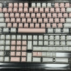 May include: Two mechanical keyboard keycap sets, one with pink and white keys, the other with white and pink keys. The keycaps are arranged in a standard QWERTY layout, packaged in a clear plastic tray.