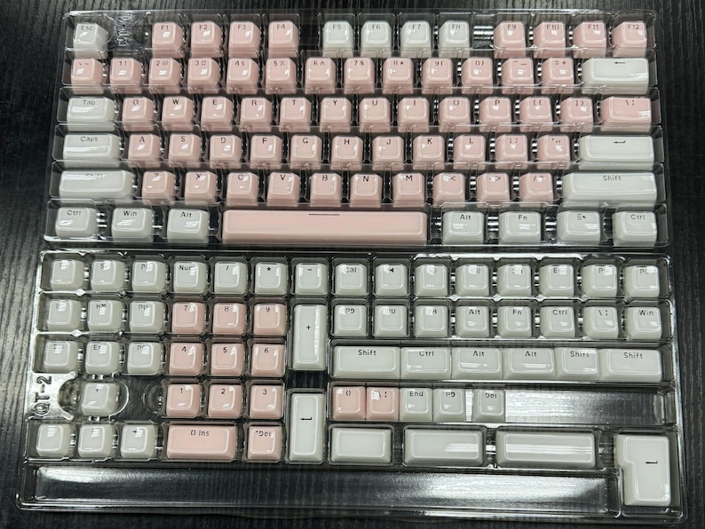 May include: Two mechanical keyboard keycap sets, one with pink and white keys, the other with white and pink keys. The keycaps are arranged in a standard QWERTY layout, packaged in a clear plastic tray.