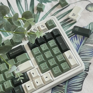 Puede incluir: Un teclado mecánico con teclas en tonos verde salvia, verde bosque oscuro y blanco hueso. Varias teclas muestran un pequeño icono de rama de olivo. El teclado numérico y las teclas de flecha son visibles con letras negras estándar.