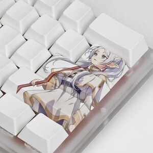 May include: A custom keyboard keycap featuring an anime-style illustration of a character with long white hair, wearing a white and gold outfit, and holding a staff. The keycap is surrounded by white keys.