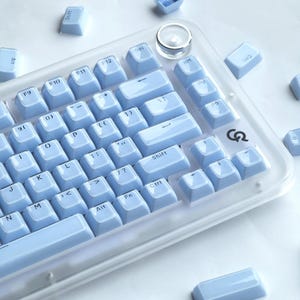 以下が含まれることがあります： 透明なフレームのライトブルーのメカニカルキーボード。キーボードは、白い文字が入ったライトブルーのキーキャップを備えています。いくつかの緩いキーキャップが散らばっています。キーボードの右上隅には、丸くて透明なノブがあります。