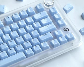 Könnte beinhalten: Eine hellblaue mechanische Tastatur mit transparentem Rahmen. Die Tastatur hat hellblaue Tastenkappen mit weißer Beschriftung. Mehrere lose Tastenkappen sind verstreut. Die Tastatur hat einen runden, transparenten Knopf oben rechts.