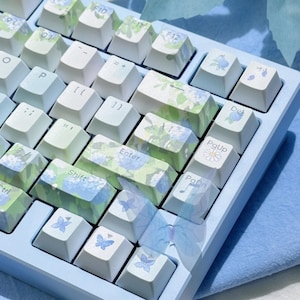 以下が含まれることがあります： 白と花のキーキャップが付いた、水色のメカニカルキーボード。キーキャップには、青と緑の花柄と蝶のデザインが施されています。キーボードは水色のフレームで、視聴者に向かって傾いています。