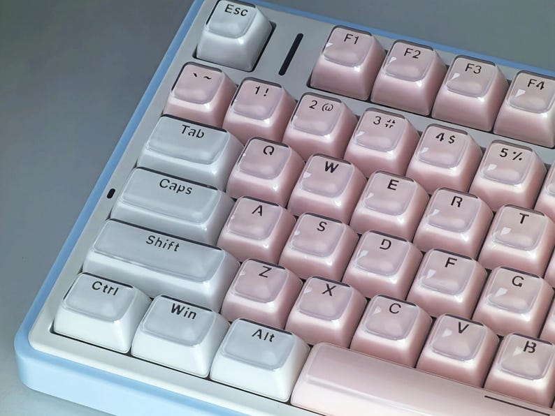 May include: Close-up of a compact mechanical keyboard with a light blue frame. The keycaps are a gradient of white to pink, with white lettering. The keyboard has a standard QWERTY layout with function keys.