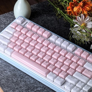 May include: A light blue mechanical keyboard with white and pink keycaps. The keyboard has a compact layout with a variety of function keys. The keycaps are a soft pink color, and the keyboard has a light blue frame.