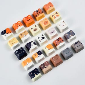 K&ouml;nnte beinhalten: Eine Sammlung von Tastenkappen mit Katzenmotiven in verschiedenen Farben, darunter Orange, Schwarz, Wei&szlig; und Braun. Jede Tastenkappe zeigt ein anderes Katzengesicht-Design. Die Tastenkappen sind in ordentlichen Reihen und Spalten angeordnet.